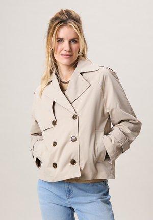 Vrouw met blond haar die een beige dubbelgeknoopt jasje en lichtblauwe spijkerbroek draagt, staand met handen in de zakken tegen een effen achtergrond.