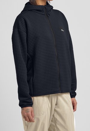 Person iført en sort quiltet hættetrøje med lynlås og lyse beige bukser, med synlige tatoveringer på venstre hånd mod grå baggrund.