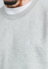 Sudadera gris clara con un tejido texturizado, que cuenta con un cuello redondo acanalado y un pequeño logo bordado en blanco en el lado izquierdo del pecho.