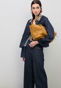 Veste en denim avec col imprimé léopard, associée à un jean. Le mannequin tient un grand sac à main texturé jaune moutarde. Boucles d'oreilles rondes rouges mises en avant.