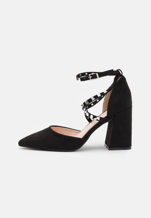 Czarny zamszowy but na wysokim obcasie z zaostrzonym noskiem i masywnym obcasem. Posiada dwie paski wokół kostki z srebrnymi ćwiekami oraz miękkie, teksturowane wykończenie.