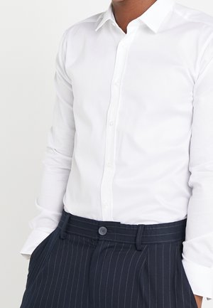 Mężczyzna w dopasowanej, białej koszuli zapinanej na guziki, włożonej do granatowych spodni w prążki, z rękami w kieszeniach.