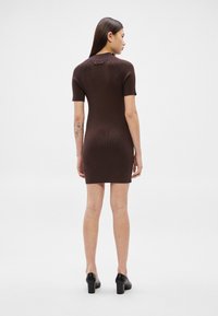 Γυναίκα με μακριά σκούρα μαλλιά, φορώντας στενό σκούρο καφέ ριγέ φόρεμα με κοντά μανίκια και μαύρα παπούτσια με τακούνι, στέκεται με την πλάτη προς τα εμπρός σε απλό φόντο.