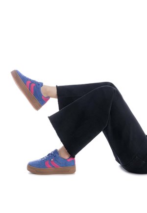 Zapatillas de ante azules con rayas rosas, suela de goma marrón y diseño de cordones. Llevadas con unos vaqueros amplios negros con dobladillos deshilachados.