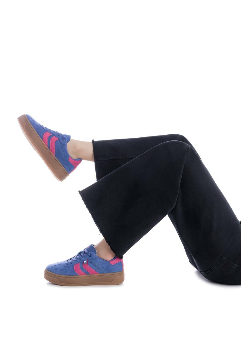 Zapatillas de ante azules con rayas rosas, suela de goma marrón y diseño de cordones. Llevadas con unos vaqueros amplios negros con dobladillos deshilachados.