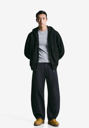 Schwarze Zip-up Kapuzenjacke über einem grauen Shirt, locker sitzende dunkelblaue Hose und beige Schuhe; schlichtes Design mit einer entspannten Silhouette.
