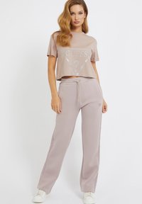 Svetlá taupe skrátená tričko s strieborným logom "GUESS" a zodpovedajúcimi vysokými joggers, vyrobené z jemnej látky, s elastickým pásom a vreckami.