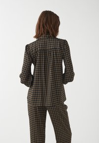 Schwarz-blusiges Oberteil mit goldenem geometrischem Muster, das puffartige Ärmel und ein gefaltetes Rückendesign aufweist. Kombiniert mit passenden, gemusterten Hosen.