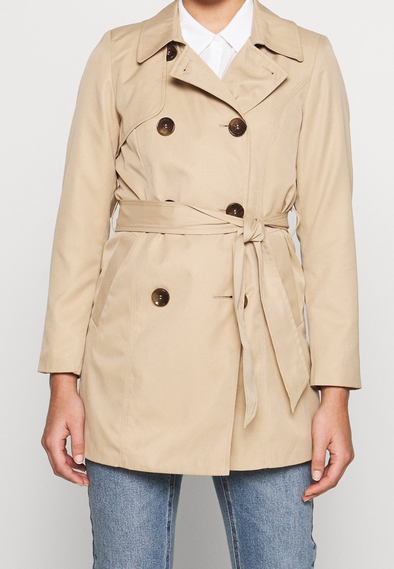 Trench-coat beige à double boutonnage avec ceinture nouée à la taille, porté sur une chemise blanche et un jean bleu, vu du cou jusqu'aux hanches.