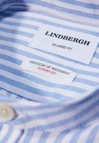Colletto di camicia a righe blu e bianche con etichette che mostrano "Lindbergh Relaxed Fit" e "Freedom of Movement Superflex." Materiale in morbido cotone.