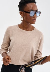 Beżowy sweter dzianinowy z okrągłym dekoltem i długimi rękawami, w zestawieniu z dużymi okularami w stylu żółwi i wzorzystym czarnym szalem.