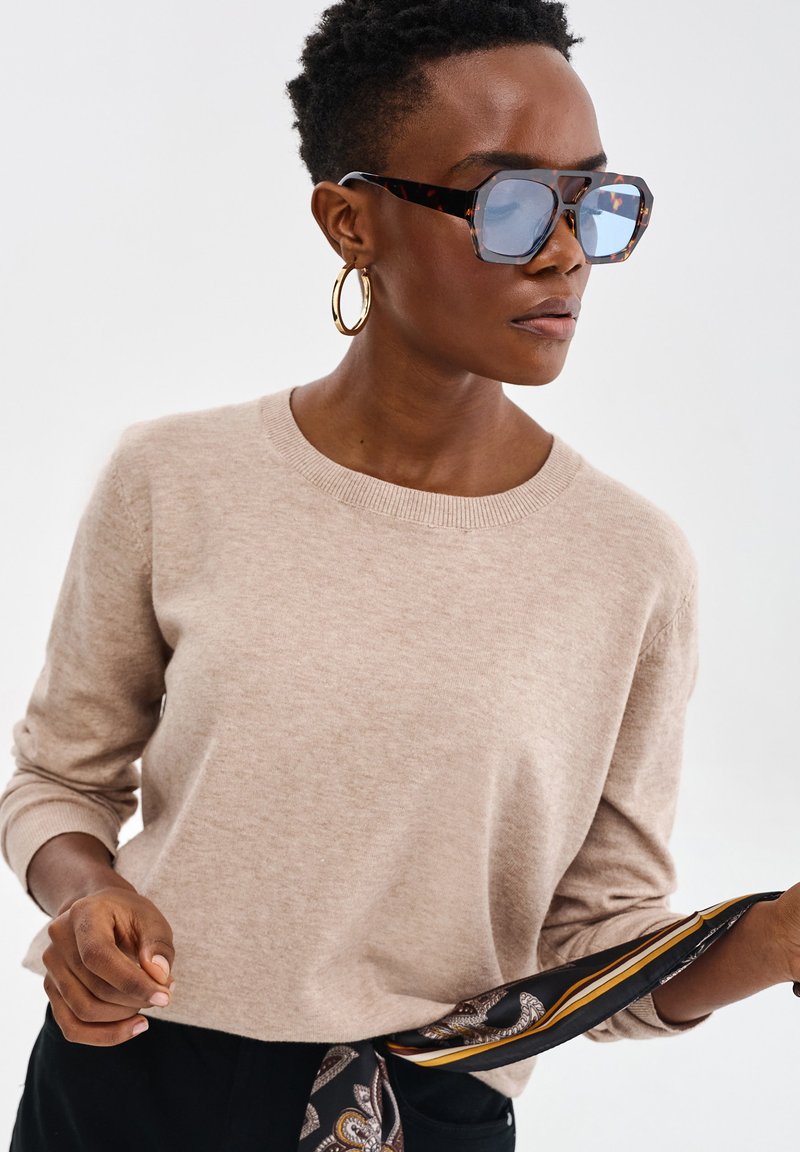 Beżowy sweter dzianinowy z okrągłym dekoltem i długimi rękawami, w zestawieniu z dużymi okularami w stylu żółwi i wzorzystym czarnym szalem.