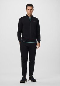 Muškarac stoji odjeven u crnu jaknu na zatvarač, crne hlače, crne tenisice i teal majicu ispod, a pozadina je jednostavna svijetla.