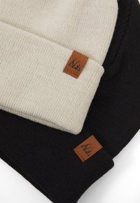 Due due berrettini in maglia, uno crema e uno nero, ciascuno con un'etichetta in pelle marrone con un design di montagna. Texture morbida, design semplice.