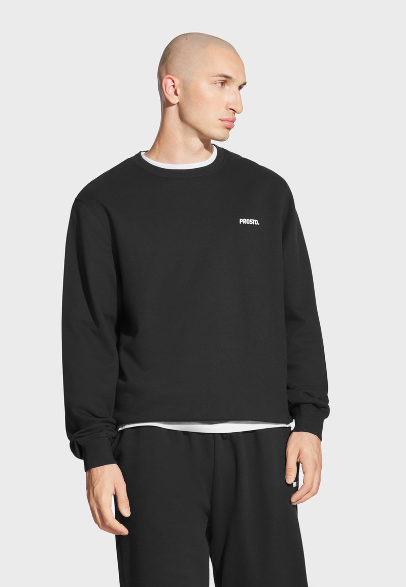 Czarna bluza z okrągłym dekoltem i długimi rękawami, z białym logo "PROSTO." na lewej piersi, w zestawie z czarnymi spodniami.