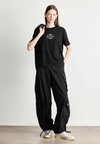 Zwarte katoenen T-shirt met een wit logo, gecombineerd met losse zwarte cargo broeken met zakken en ritsen. Outfit afgemaakt met grijze en beige sneakers.