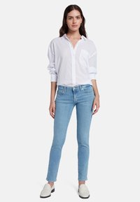 Camicia bianca a maniche lunghe con bottoni e tasca, abbinata a jeans skinny azzurri chiari e scarpe slip-on color avorio. Postura dritta.
