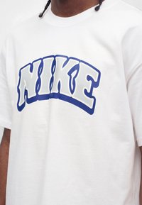 Άτομο που φοράει λευκό T-shirt με καμπυλωτό, έντονο λογότυπο "NIKE" σε λευκά γράμματα με μπλε περίγραμμα στο στήθος.