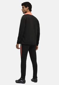 Tuta sportiva nera con colletto alto, caratterizzata da dettagli corallo, design aderente e gambe slim. Realizzata in materiale morbido e flessibile.