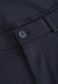 Tejido navy con una textura suave, que presenta un cierre de botón y una cinturilla elástica. Incluye detalles de costura vertical para dar estructura.