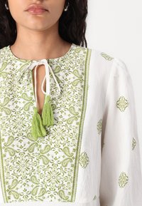 Hvit bluse med grønn paisley- og blomsterbroderi, rund hals med knyting og dusk, samt lange puffermer.