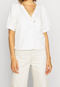 Blouse en coton blanc avec col en V, manches courtes bouffantes, deux poches avant et fermeture par boutons. Texture lisse, coupe décontractée, design léger.