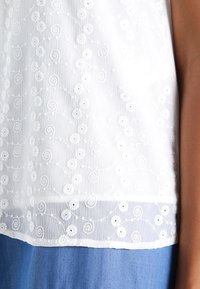 Haut blanc brodé avec un motif floral et des accents en spirale, texture transparente et un ourlet inférieur uni, associé à un tissu en denim.