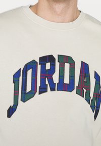 Felpa beige chiaro con la scritta "JORDAN" in grandi lettere ricamate, con un motivo a scacchi blu, verde e rosso. Collo rotondo.
