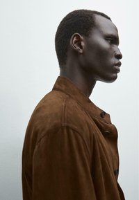 Veste en daim marron avec un col haut et une fermeture à boutons, mise en valeur par une texture douce et une coupe ajustée. Vue de profil d'un mannequin.