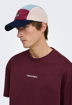 Homme portant une casquette color block bleu, bordeaux et beige et un t-shirt bordeaux avec le texte "Leave blank" sur la poitrine, tourné vers la gauche.