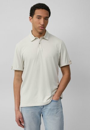 Helles beige Poloshirt mit Kragen und zwei Knöpfen. Kurze Ärmel mit umgeschlagenen Bündchen, kombiniert mit hellblauen Jeans. Glatte Stofftextur.