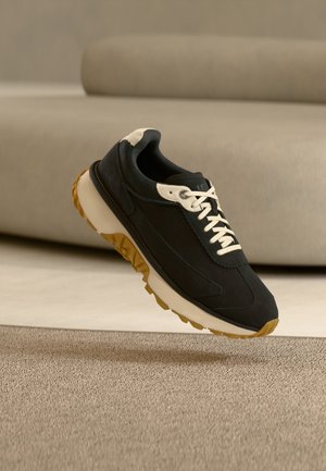 Baskets noir et blanc à semelle en gomme flottant au-dessus d’un tapis beige dans une pièce aux tons neutres avec un canapé beige courbé en arrière-plan.