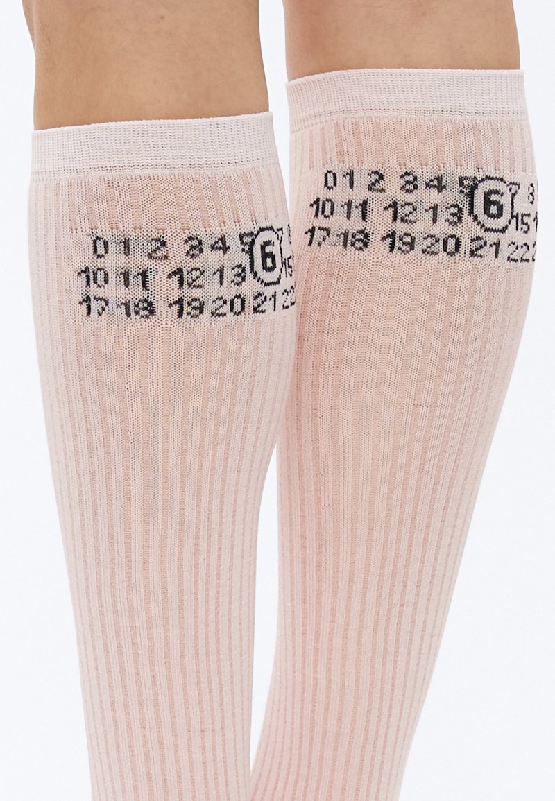 Knæhøje, ribstrikkede strømper i blød rosa med sort taldesign og logo trykt nær toppen. Materialet virker strækbart og let.