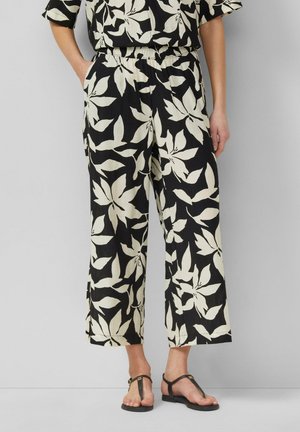 Zwarte cropped broek met grote witte bloemenprint, elastische tailleband, zijzakken, gedragen met zwarte T-strap sandalen op een effen achtergrond.