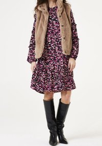 Kobieta w różowo-czarną wzorzystą sukienkę, beżową kamizelkę z sztucznego futra i wysokie czarne botki, stojąca na białym tle.