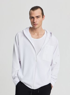 Giovane uomo con capelli pettinati all’indietro che indossa una felpa bianca con zip sopra una canottiera bianca, mani nelle tasche dei pantaloni neri, sfondo neutro.