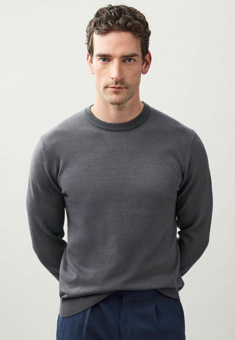 Suéter de punto gris con cuello redondo, puños y dobladillo acanalados. El material tiene una textura suave y un diseño ajustado, combinado con pantalones de color azul oscuro.
