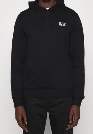 Homme portant un sweat à capuche noir avec poche avant et logo blanc EA7 Emporio Armani sur la poitrine gauche, assorti à un pantalon noir.