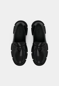 Black loafers with a smooth leather upper, rounded shape, white rubber soles, and a decorative metal buckle accent.