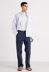 Lichtblauwe overhemden met knopen, een wit T-shirt eronder, marineblauwe cargobroek en bruine sandalen. Het shirt heeft een borstzakje; de broek heeft meerdere zakken.