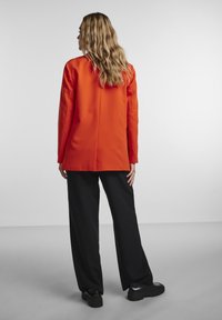 Femme aux longs cheveux ondulés portant un blazer orange, un pantalon noir à jambes larges et des chaussures plateformes noires, debout de dos sur un fond uni.