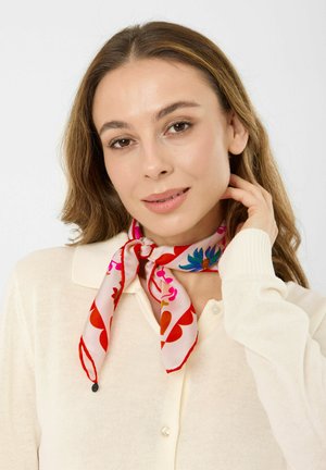 Frau mit hellbraunen Haaren, die einen cremefarbenen, geknöpften Pullover und einen bunten, floralen Seidenschal um den Hals trägt und ihr Haar berührt.