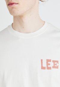 T-shirt en coton blanc avec un imprimé "LEE" en rouge corail à effet usé sur le devant. Encolure arrondie, manches courtes, texture lisse.