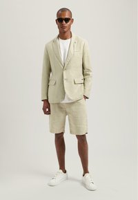 Hellgrüner Leinenblazer, weißes T-Shirt, passende Shorts und weiße Sneakers. Schwarze Sonnenbrille und ein kleines Reverspin-Detail.