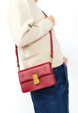 Sac bandoulière en cuir rouge de forme rectangulaire, avec des accents métalliques dorés et un fermoir texturé, porté sur un pull beige et un jean bleu.