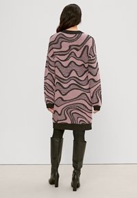 Robe-pull tricotée à motifs roses et noirs, manches longues, col ras du cou et ourlet côtelé. Portée sur un legging noir et des bottes à talons hauts.