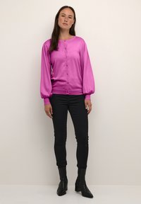 Rosa Button-Up-Strickjacke mit langen, gepufften Ärmeln, kombiniert mit schwarzen, figurbetonten Jeans und schwarzen Knöchelstiefeln. Glatte Textur und lässiges Design.