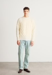 Krém színű, kötött pulóver bordázott mandzsettákkal, világoskék farmerrel és sötétbarna cipővel párosítva. Egyszerű, klasszikus dizájn és kényelmes szabás.