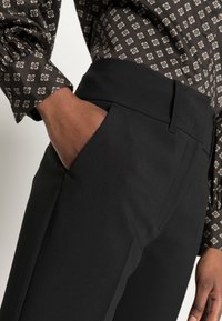 Svarta skräddarsydda byxor med en slät yta, utrustade med sidofickor och hög midja, ihop med en mönstrad brun skjorta.