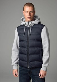 Doudoune marine sans manches avec un hoodie gris à manches longues. Présente un design matelassé, une fermeture éclair et des poignets côtelés gris. Portée sur un jean en denim foncé.